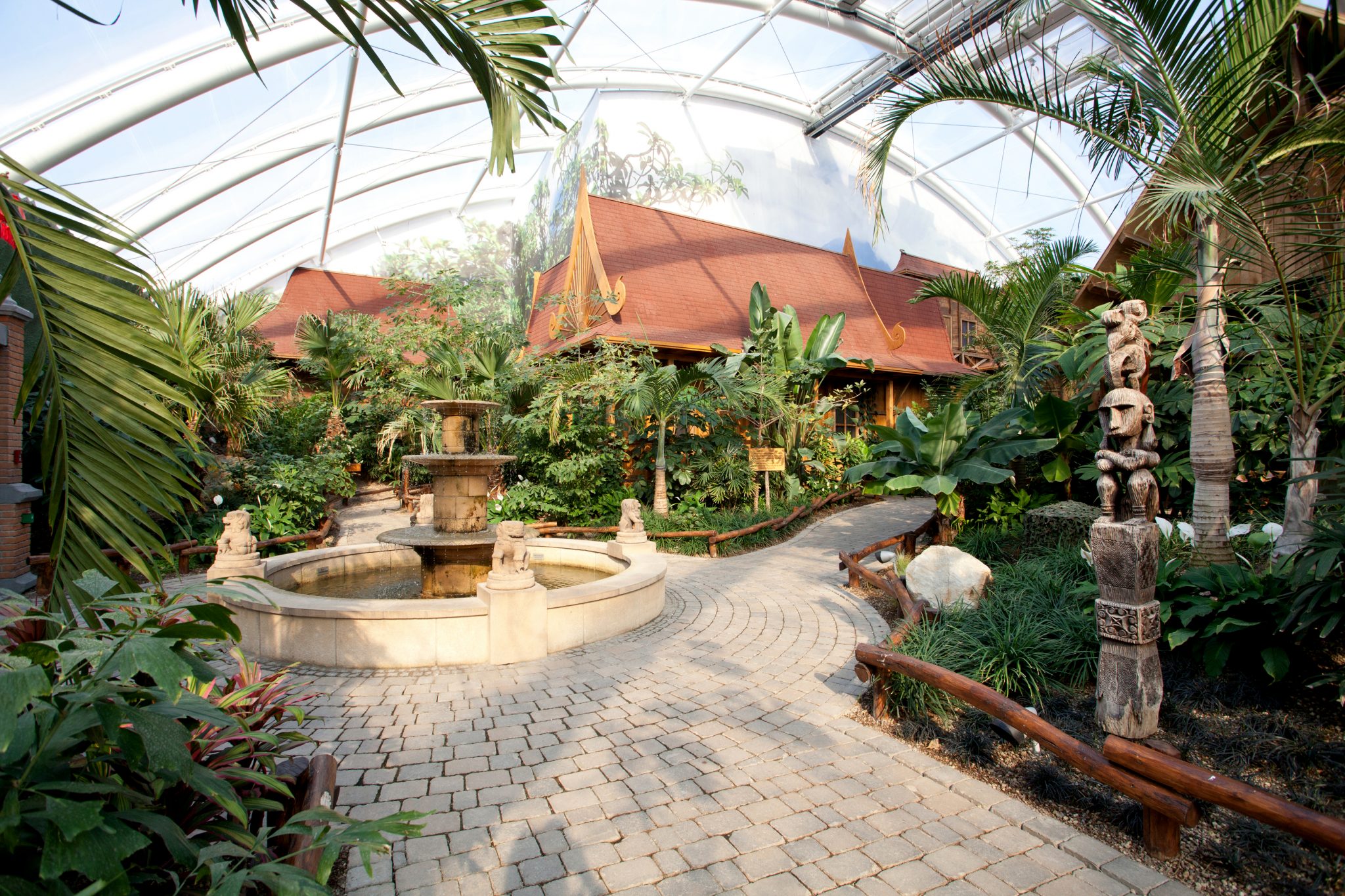 GBMpic_ Ein tropisches Gewächshaus mit einem zentralen Brunnen, üppiger Vegetation und einem gewundenen Pfad, der zu einem Pavillon im thailändischen Stil führt.