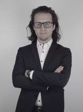 GBMpic_ Ein Mann im schwarzen Anzug und mit Brille steht mit verschränkten Armen vor einem schlichten Hintergrund.