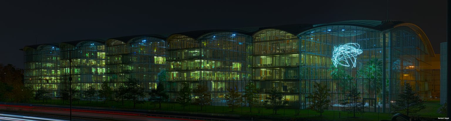 GBMpic_ Ein nächtlicher Panoramablick auf ein modernes Gebäude mit beleuchteter Glasfassade und einer markanten Lichtprojektion an der Außenseite.