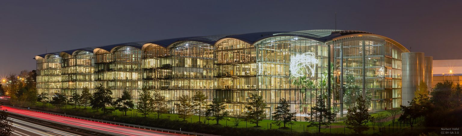 GBMpic_ Modernes Glasgebäude mit abgerundeten Abschnitten, nachts beleuchtet, mit Lichtspuren vom vorbeifahrenden Verkehr im Vordergrund.