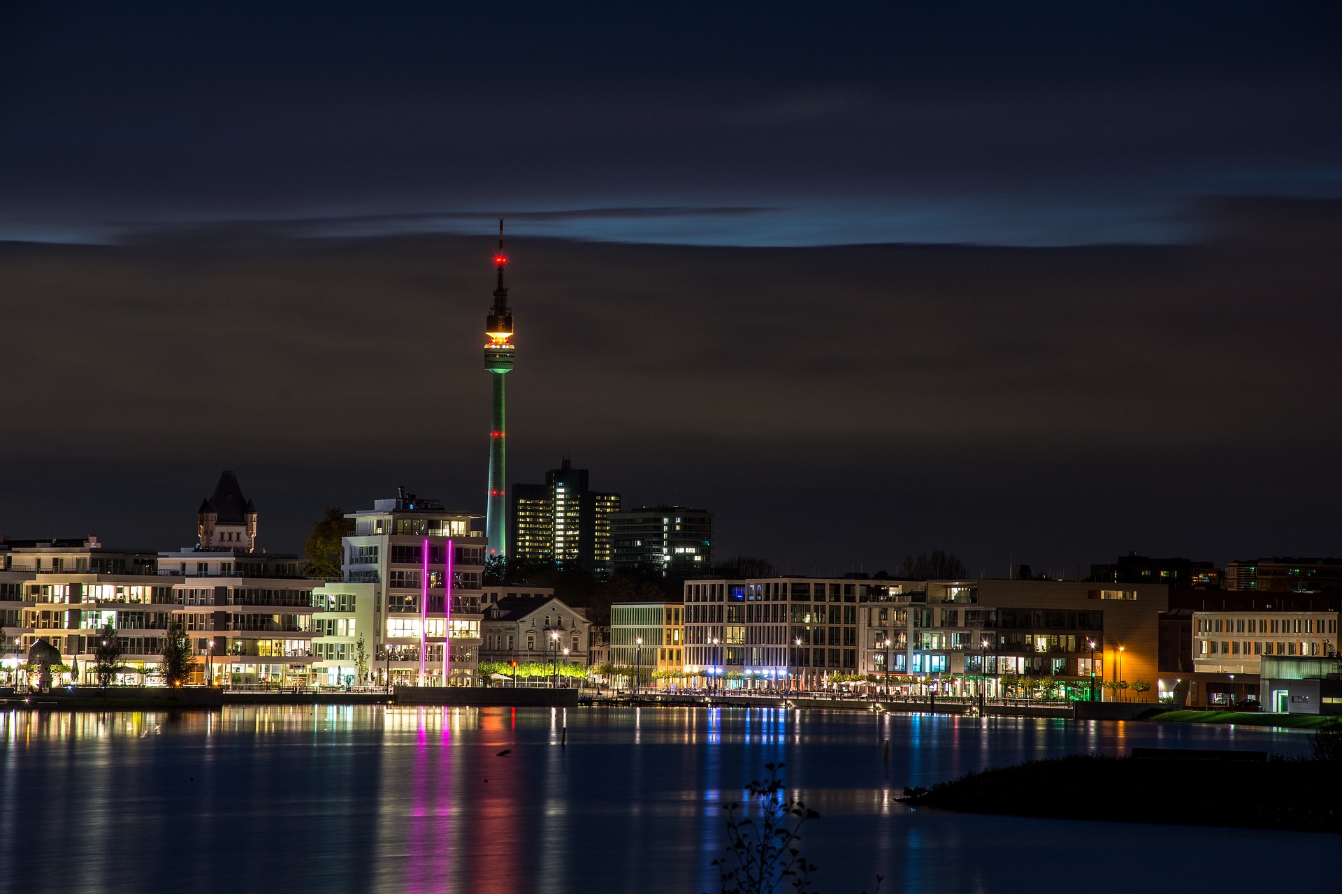 GBMpic_ Skyline der Stadt bei Nacht mit einem beleuchteten Turm, der sich im Wasser spiegelt.