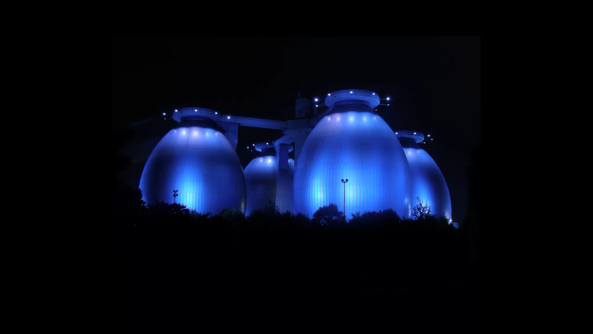 GBMpic_ Blau beleuchtete kuppelförmige Strukturen bei Nacht, mit schwarzem Hintergrund und Baumsilhouetten im Vordergrund.