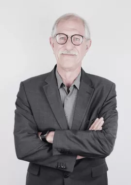 GBMpic_ Rainer Reimers - Ein Mann mit weißem Haar und Schnurrbart, Brille und grauem Anzug steht mit verschränkten Armen vor einem schlichten weißen Hintergrund.