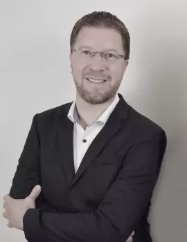 GBMpic_ Marco Podwojewski Ein Mann mit kurzen Haaren, Brille und Bart lächelt, trägt einen dunklen Anzug, ein weißes Hemd und verschränkt die Arme. Er steht vor einem schlichten hellen Hintergrund.
