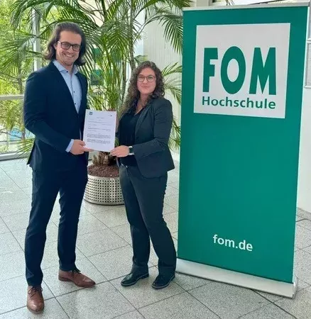 GBMpic_ Zwei Personen stehen drinnen neben einem Banner mit der Aufschrift „FOM Hochschule“ und halten ein Dokument zusammen. Im Hintergrund sind eine große Topfpflanze und Fenster zu sehen, die die Zusammenarbeit im dualen Studium für den Studiengang Bachelor of Engineering – Bauingenieurswesen symbolisieren.