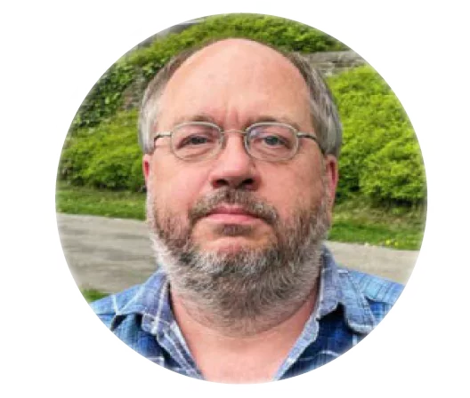 GBMpic_ Ein Mann mittleren Alters mit Brille, Bart und kariertem Hemd steht im Freien, mit Grünflächen und einem Weg im Hintergrund, und denkt über seine Erfahrungen im Projektmanagement für Airport 2000 Plus nach.