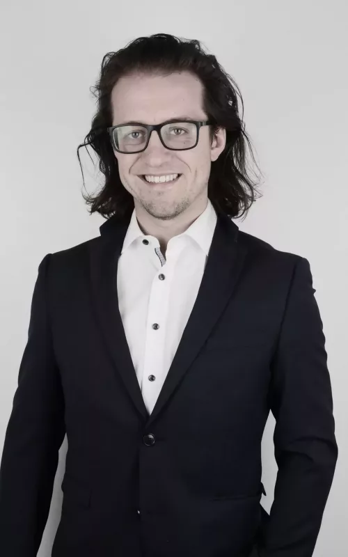 GBMpic_ Eine Person mit langen Haaren und Brille, die einen dunklen Anzug und ein weißes Hemd trägt, lächelt vor einem schlichten Hintergrund in die Kamera.