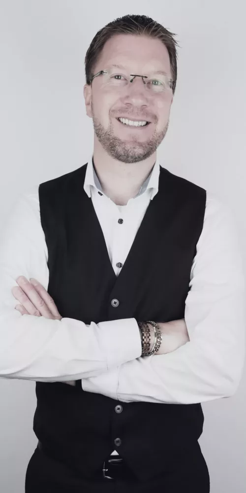GBMpic_ Eine Person mit Brille, weißem Hemd und schwarzer Weste steht lächelnd mit verschränkten Armen vor einem schlichten Hintergrund.